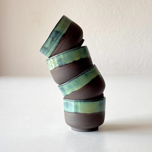 May include: A stack of four small ceramic cups with brown and green glaze. The cups are stacked in a leaning tower shape.