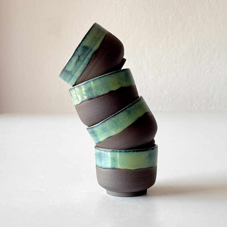 May include: A stack of four small ceramic cups with brown and green glaze. The cups are stacked in a leaning tower shape.