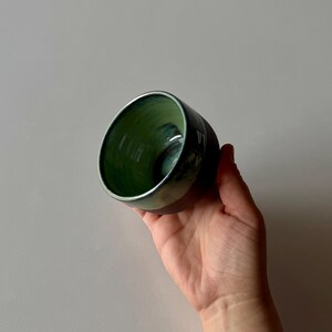 May include: A small, green and black ceramic cup with a glazed finish. The cup is held in a hand.
