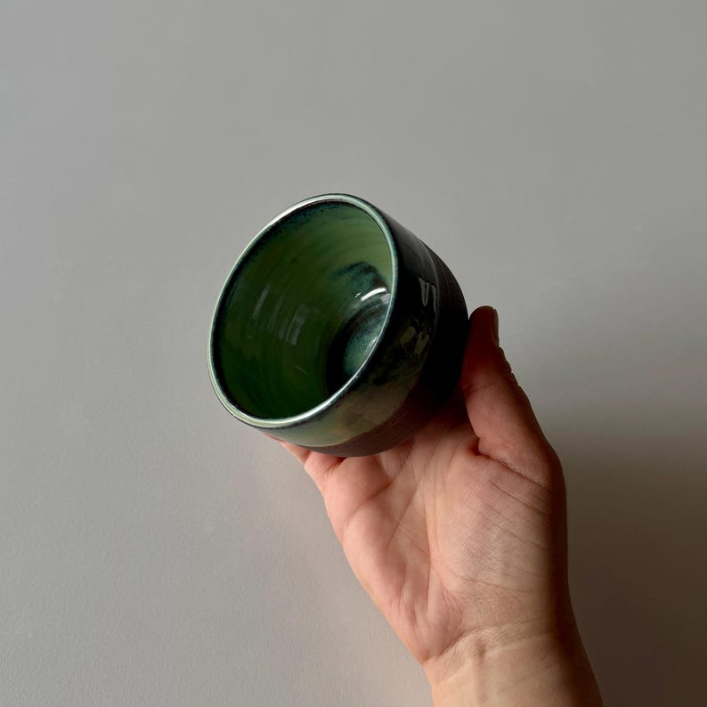 May include: A small, green and black ceramic cup with a glazed finish. The cup is held in a hand.