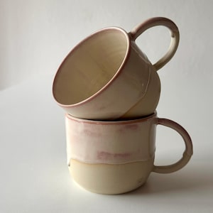 Pode incluir: Duas canecas de cerâmica feitas à mão empilhadas uma sobre a outra. As canecas são de cor bege creme com uma faixa rosa-avermelhada no meio e na borda. Cada caneca tem uma alça arredondada. As canecas estão sobre uma superfície branca.