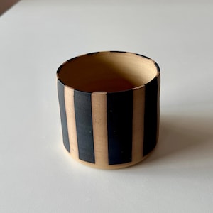 May include: A cylindrical ceramic cup with a beige interior and exterior. The exterior features vertical black stripes alternating with beige stripes. The cup is sitting on a white surface.