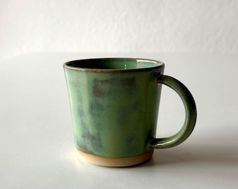 May include: A green ceramic mug with a brown rim and a slightly flared shape. The mug has a unique, mottled glaze that gives it a rustic look.