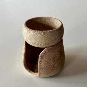 K&ouml;nnte beinhalten: Ein zweiteiliger Keramik-&Ouml;lbrenner mit einer rauen, sandigen, beigefarbenen Textur. Der obere Teil ist eine kreisf&ouml;rmige Schale f&uuml;r &Ouml;l, w&auml;hrend der untere Sockel eine breite, bogenf&ouml;rmige &Ouml;ffnung aufweist, um eine W&auml;rmequelle aufzunehmen.