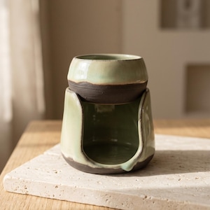 May include: A two-piece ceramic wax warmer featuring a dark brown base with a smooth, light sage green glaze on the upper sections. The top bowl sits securely on the arched stand, which has a wide front opening for a tea light candle.