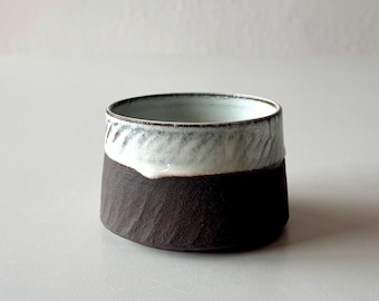 May include: A small, round ceramic bowl with a dark brown exterior and a white interior. The bowl has a textured, slightly uneven surface and a white band around the top.