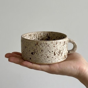 Könnte beinhalten: Eine gesprenkelte Keramiktasse mit einem abgerundeten Henkel. Die Tasse hat eine cremefarbene Basis mit braunen Sprenkeln. Die Tasse wird in der Hand einer Person gehalten. Die Tasse hat eine neutrale Farbe.