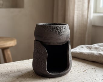Puede incluir: Un calentador de cera de cerámica gris carbón oscuro con una superficie texturizada y craterizada que recuerda a la luna. El objeto consta de una sección superior en forma de cuenco sobre una base con una gran abertura arqueada.