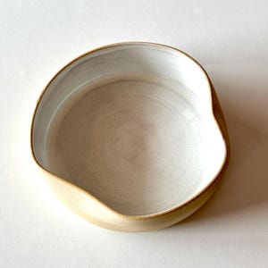 Pode incluir: Uma tigela de cerâmica branca com uma borda bege. A tigela tem uma forma ligeiramente irregular e um interior liso e esmaltado.
