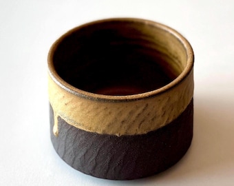May include: A cylindrical ceramic cup featuring a two-tone design with a dark brown, textured lower section and a lighter, tan-colored upper rim. The tan glaze drips slightly down the side, creating a distinct contrast against the dark base.
