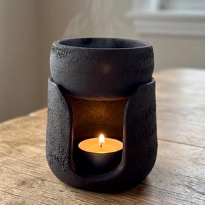 Op de afbeelding: Zwarte, getextureerde keramische etherische olie diffuser met een brandende kaars erin. Stoom stijgt op van de bovenkant van de diffuser, die op een houten oppervlak staat. Het ontwerp heeft een afgeronde bovenkant en een gebogen opening voor de kaars.