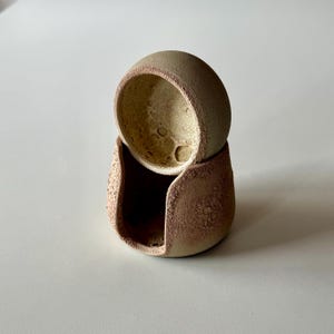 K&ouml;nnte beinhalten: Ein zweiteiliges Keramikobjekt, bestehend aus einer abgerundeten, strukturierten beigen Schale, die auf einer gr&ouml;&szlig;eren, dunkelbraunen Basis mit einer breiten, bogenf&ouml;rmigen &Ouml;ffnung ruht. Die Au&szlig;enfl&auml;chen haben eine raue, k&ouml;rnige Oberfl&auml;che, w&auml;hrend das Innere der oberen Schale eine hellere, gesprenkelte und por&ouml;se Textur aufweist.