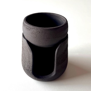 May include: A black ceramic oil burner with a textured finish. The burner has a small bowl on top and a larger base with a cutout for a tea light.