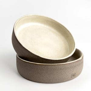 May include: Two ceramic bowls stacked on top of each other. The bottom bowl is a speckled, dark brown color, and the top bowl is a lighter beige color. The bowls have a simple, modern design and are likely handmade.