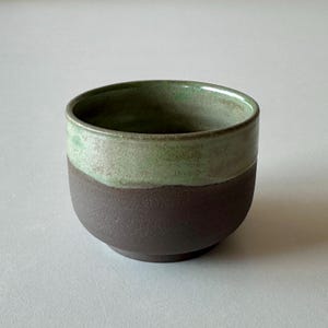 Puede incluir: Un cuenco de cerámica marrón oscuro con un esmalte verde claro en la mitad superior. El cuenco tiene una forma ligeramente redondeada y una superficie texturizada.
