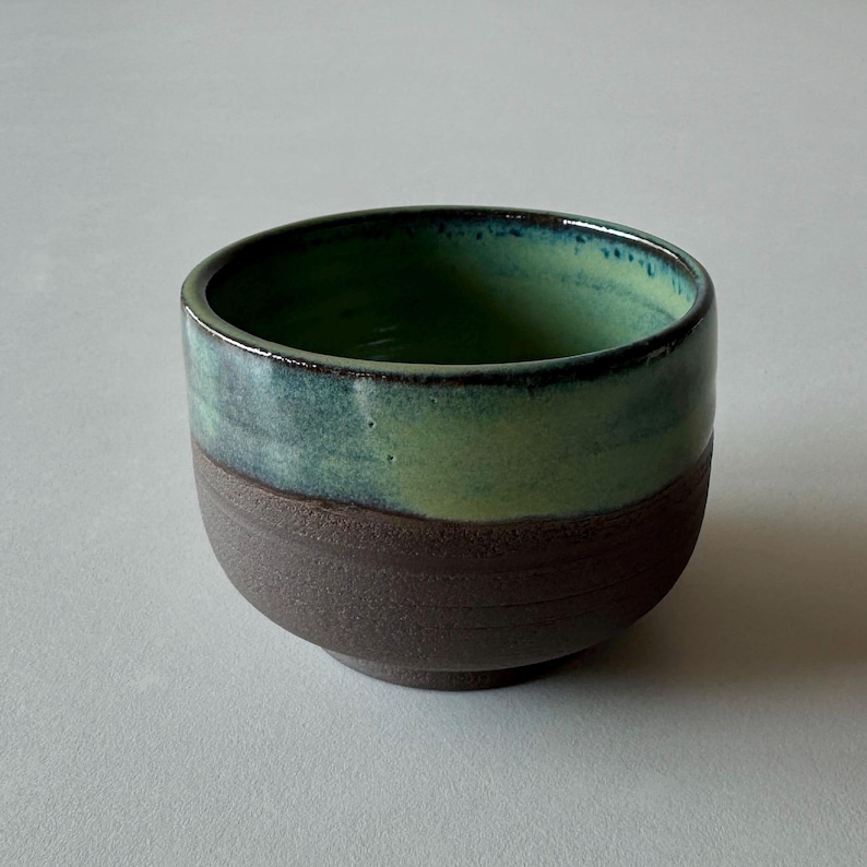 May include: A small, green and brown ceramic bowl with a textured bottom. The bowl is glazed in a light green color with a darker green rim.