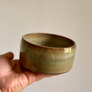 Op de afbeelding: Een handgemaakte keramische kom met een unieke, asymmetrische vorm. De kom heeft een glanzend, gespikkeld glazuur in groen- en bruintinten, met een donkerdere rand. De kom wordt in een hand gehouden, wat de grootte en vorm laat zien.
