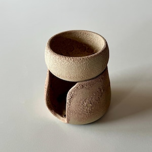 May include: A two-part ceramic vessel with a textured surface. The top is a small bowl, stacked above a rounded base with a curved opening. The colors are warm beige and reddish-brown. The piece is likely handmade.