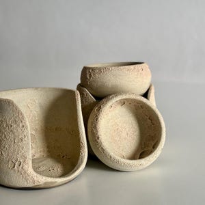 K&ouml;nnte beinhalten: Eine Ansammlung von drei Keramikgef&auml;&szlig;en mit rauer, kraterartiger Textur in blassem Beige. Ein Gef&auml;&szlig; ist seitlich offen und geschwungen, w&auml;hrend die anderen beiden kleine, runde Schalen sind, die alle eine por&ouml;se, mond&auml;hnliche Oberfl&auml;che aufweisen.
