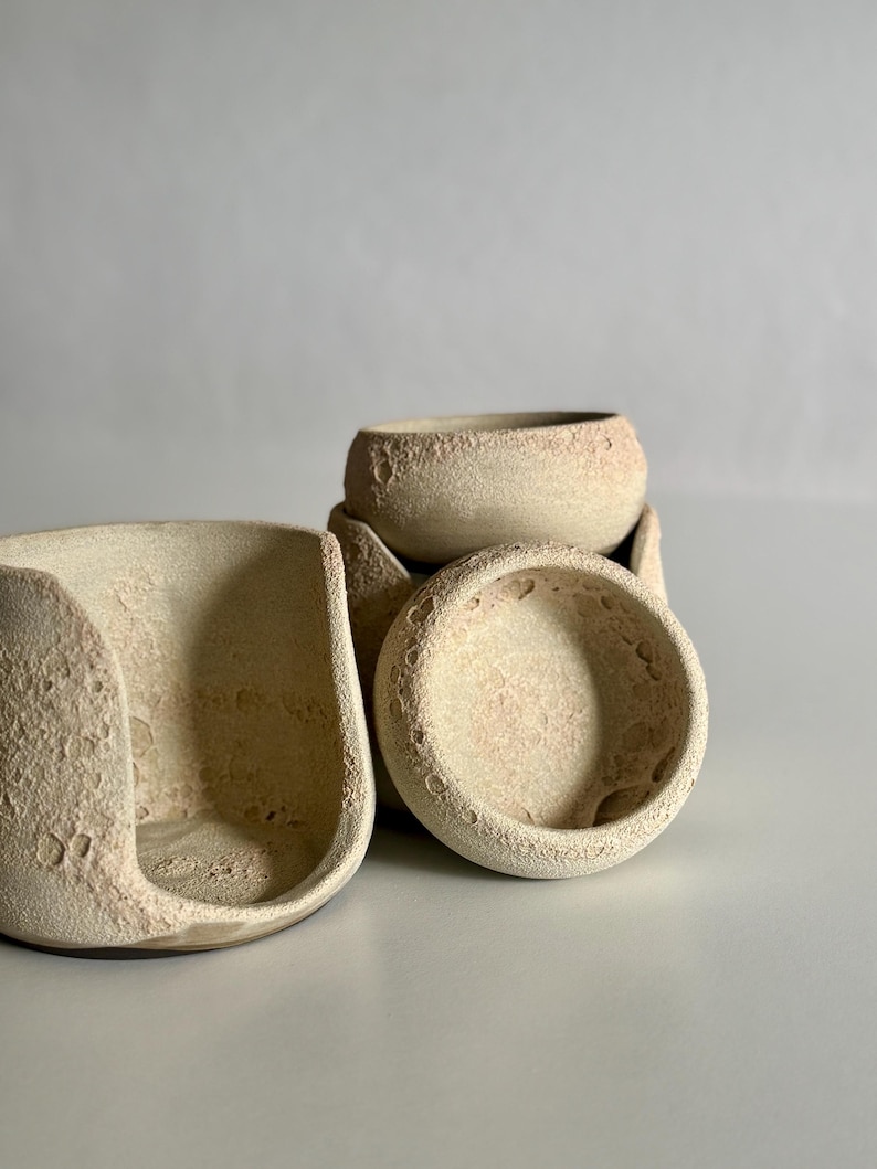 K&ouml;nnte beinhalten: Eine Ansammlung von drei Keramikgef&auml;&szlig;en mit rauer, kraterartiger Textur in blassem Beige. Ein Gef&auml;&szlig; ist seitlich offen und geschwungen, w&auml;hrend die anderen beiden kleine, runde Schalen sind, die alle eine por&ouml;se, mond&auml;hnliche Oberfl&auml;che aufweisen.