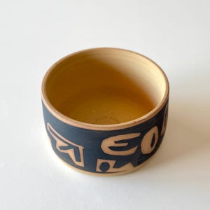 May include: A ceramic bowl with a dark blue band decorated with abstract shapes and letters. The interior of the bowl is a light tan colour. The bowl has a round shape and a simple, modern design.