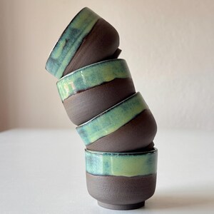 May include: A stack of four small ceramic cups with a dark brown base and a light green glaze. The cups are stacked in a leaning tower shape.