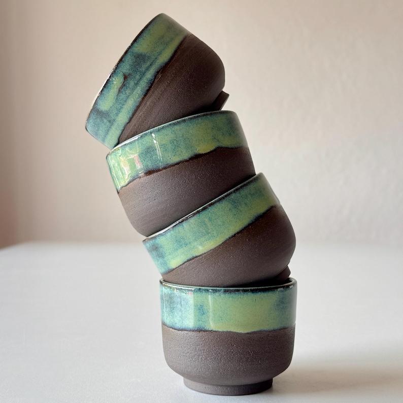 May include: A stack of four small ceramic cups with a dark brown base and a light green glaze. The cups are stacked in a leaning tower shape.