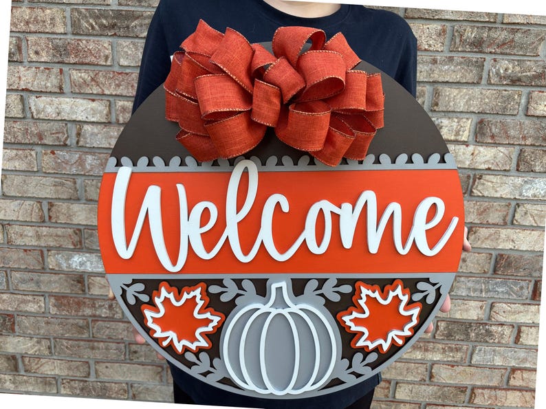 A round wooden welcome sign with a large orange bow on top, featuring the word “Welcome” in white script, a gray pumpkin outlined in white, two orange maple leaves outlined in white, and gray decorative accents.