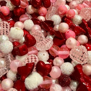 May include: A close-up of a collection of beads in various shapes and sizes. The beads are in shades of red, pink, and white, including hearts, spheres, and bow shapes. Some beads have a textured or glittery finish.