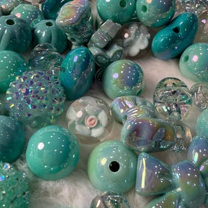 May include: A collection of various teal and iridescent beads. The beads are in different shapes and sizes, including round, oval, and faceted. Some beads have a glossy finish, while others are more matte. The beads are scattered on a white, fluffy surface.