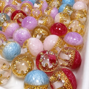 Op de afbeelding: Een verzameling kleurrijke, ronde kralen in verschillende tinten roze, blauw, rood en goud. Elke kraal heeft een gouden kroonontwerp aan de basis. De doorschijnende kralen laten licht door en zijn dicht op elkaar geplaatst.