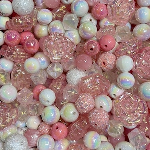 May include: A collection of assorted craft beads in shades of pink and white. The assortment includes translucent rose-shaped beads, iridescent spherical beads, faceted geometric shapes, and textured glitter-coated round beads in various sizes.