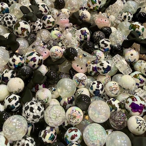 May include: A collection of various beads in white, black, and iridescent colors. Some beads have a cow print design, while others feature cartoon characters and black bow accents. The beads are of different sizes and textures.