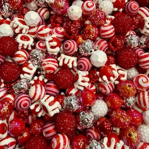May include: A close-up shot of a collection of red and white beads. The beads vary in size and shape, including round, striped, and textured designs. Some beads have a silver metallic finish, while others are solid red or white. These beads are suitable for jewelry making.