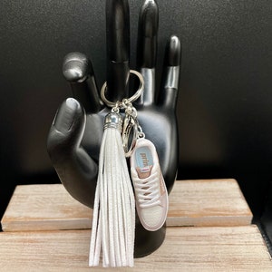 Può includere: Un portachiavi a forma di scarpa da ginnastica bianca e rosa con un pon pon bianco. Il portachiavi ha la scritta "prima" stampata sul lato della scarpa.