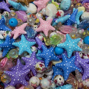 Puede incluir: Una colección de cuentas y abalorios coloridos de diversas formas y tamaños. El surtido incluye estrellas de mar, conchas marinas y cuentas redondas en tonos azules, rosas, morados y transparentes. Algunas cuentas tienen un patrón de puntos y otras son iridiscentes.