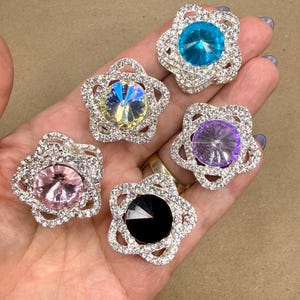 Puede incluir: Seis adornos de pedrería con forma de flor con piedras centrales de diferentes colores: azul, morado, rosa, negro, amarillo y turquesa. Los diamantes de imitación están engastados en una base de metal plateado.