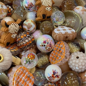 May include: A selection of colourful beads in various shapes and sizes. The beads include round, faceted, and textured designs, with a mix of glitter, floral, and animal-themed accents. Colours include brown, orange, white, and gold.
