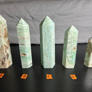 May include: Five green and brown crystal obelisks of varying sizes are arranged in a row. The crystals have a polished, faceted appearance, with natural variations in color and pattern. The obelisks stand upright against a neutral background.