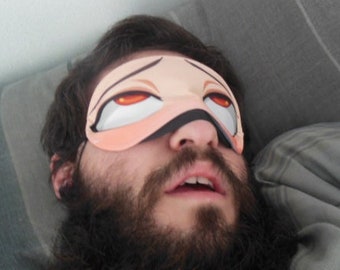 May include: A person wearing a sleep mask with a cartoon anime-style eye design. The mask is pink and white with black accents. The person has a beard and is lying down.