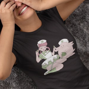 Può includere: Una maglietta grigia scura con una stampa grafica di due personaggi dei cartoni animati in abiti da infermiera che tengono foglie verdi. I personaggi sono una creatura rosa e bianca e un umano con i capelli rosa.