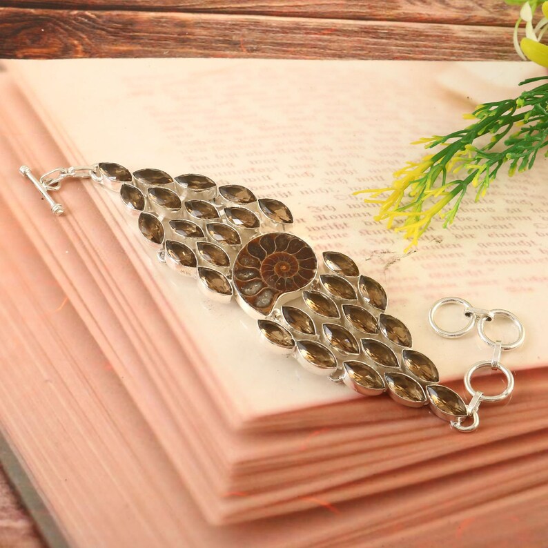 Pode incluir: Um bracelete de prata com um f&oacute;ssil de amonite castanho e detalhes de pedras preciosas castanhas. O bracelete tem um fecho de correr e um extensor de corrente.