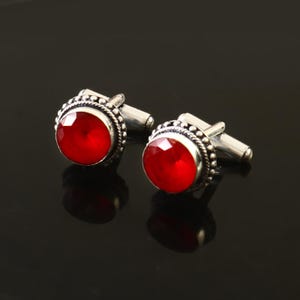Pode incluir: Um par de botões de punho de prata com detalhes de pedras preciosas vermelhas. Os botões de punho são redondos e têm uma borda de contas.