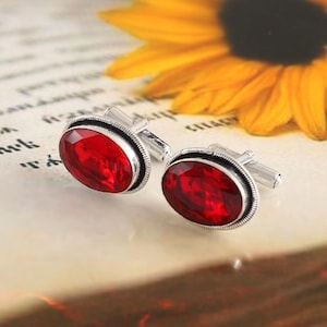 May include: A pair of silver cufflinks featuring red oval-shaped gemstones. The cufflinks have a simple design with a silver band around the gemstone.