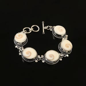 Può includere: Un bracciale in argento con sette elementi a forma di conchiglia a spirale color crema. I gusci sono incastonati in castoni d'argento e collegati da maglie d'argento. Il bracciale ha una chiusura a toggle.