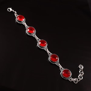 Op de afbeelding: Een zilveren armband met vijf grote, ronde, rode edelstenen. De edelstenen zijn gezet in een zilveren metalen frame en verbonden door zilveren schakels. De armband heeft een karabijnsluiting.