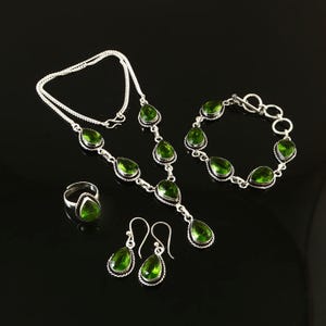 May include: A jewellery set comprising a necklace, bracelet, ring, and earrings. Each item has a silver-coloured metal setting with a teardrop-shaped, green gemstone. The set is displayed on a black surface.