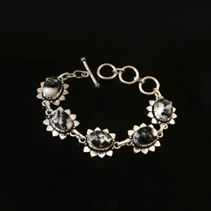 Op de afbeelding: Een zilveren armband met ovale zwart-witte stenen gezet in een bloemmotief. De armband heeft een tuimelsluiting en wordt getoond tegen een zwarte achtergrond. De stenen zijn ongeveer 2 cm lang.