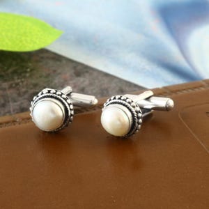 May include: A pair of silver cufflinks with white pearl accents. The cufflinks have a round shape with a beaded border.