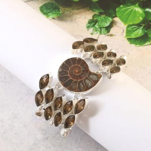 Pode incluir: Uma pulseira de prata com um f&oacute;ssil de amonite marrom e detalhes de pedras preciosas marrons.
